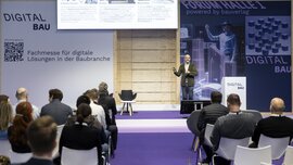 Mann steht auf einer Bühne, im Hintergrund eine lila-farbene Wand mit digitalBAU Markenabbildung, neben ihm ein Sprecherpult mit digitalBAU Logo