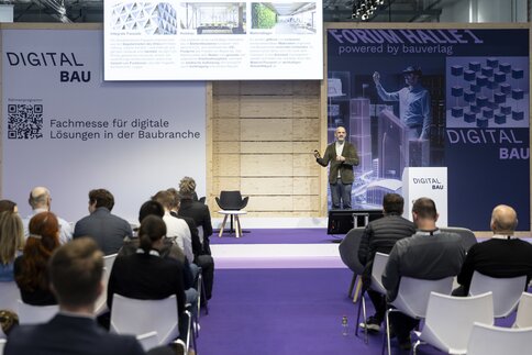 Man standing on a stage, purple wall with digitalBAU brand image in the background, lectern with digitalBAU logo next to him