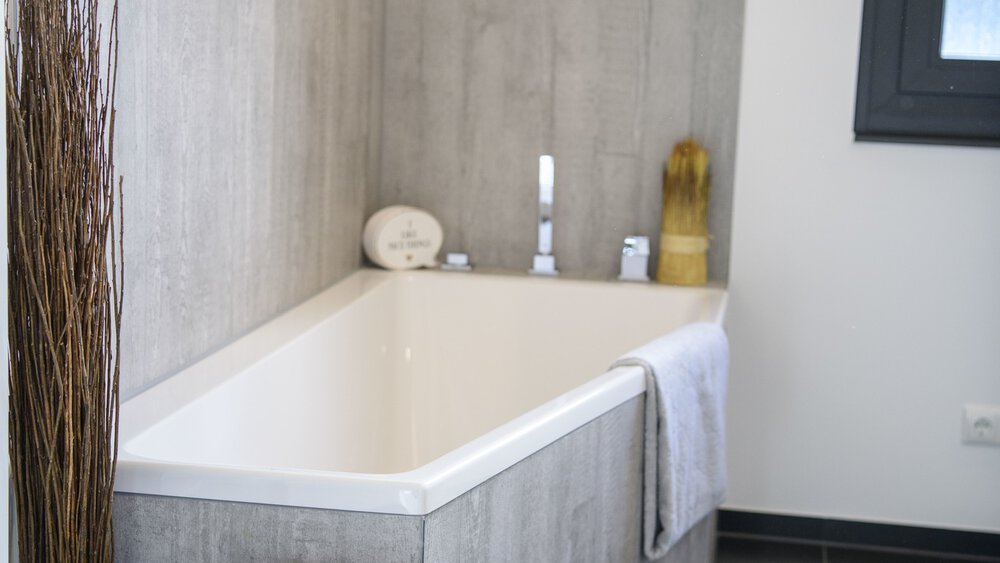 Modernes Badezimmer mit Eckbadewanne, hellgrauen Fliesen und einem über dem Rand hängenden weißen Handtuch.