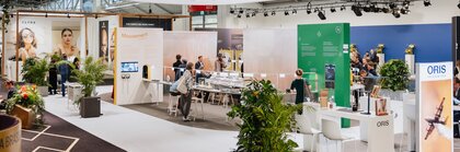 A view of several exhibition stands at INHORGENTA, featuring information boards, tables and plants, with visitors standing amongst them.