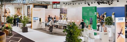 A view of several exhibition stands at INHORGENTA, featuring information boards, tables and plants, with visitors standing amongst them.