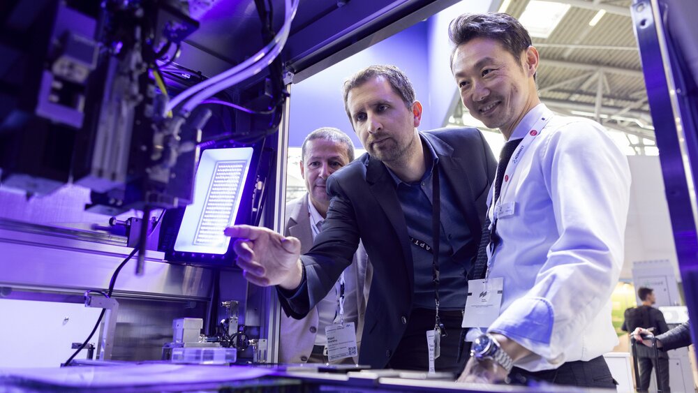 Das Innere einer Maschine mit einem steuerbaren Roboterarm. Drei Männer schauen durch eine Öffnung von außen in die Maschine hinein und lächeln.