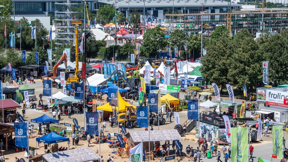 Outdoor area of a trade fair with stands, machines, flags and crowds of people on a sunny day.