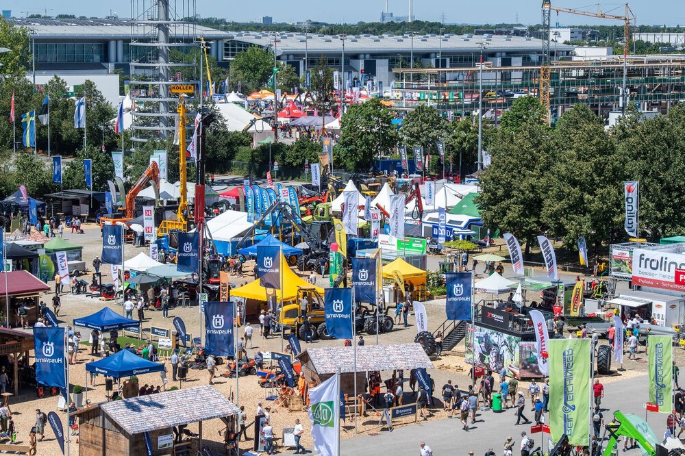 Outdoor area of a trade fair with stands, machines, flags and crowds of people on a sunny day.