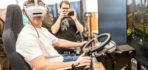Man wearing a VR headset seated at a driving simulator with a steering wheel and joystick.