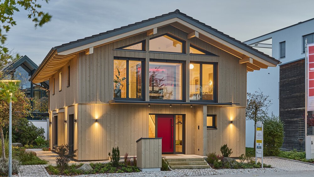 Modernes Holzhaus mit großen Fenstern, roter Tür und Außenbeleuchtung, umgeben von einem gepflegten Garten.