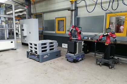 Eine Fabrikhalle mit Roboterarmen an Arbeitstischen, Paletten auf einer selbstfahrenden Plattform und Maschinen im Hintergrund.