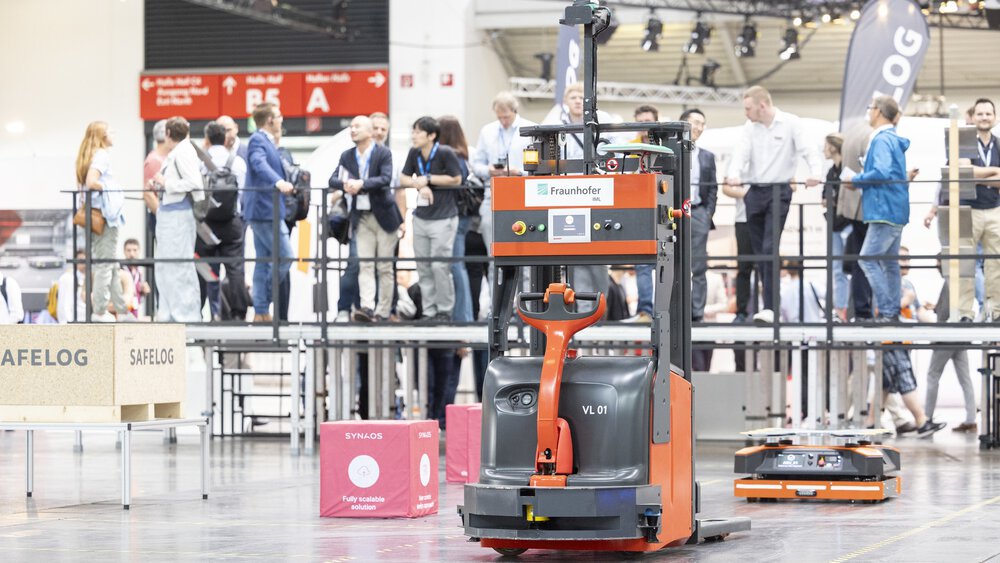 A robotized forklift truck on a demonstration area in an exhibition hall, in front of spectators on a platform.