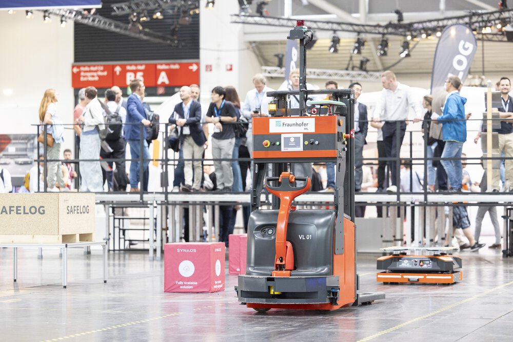 A robotized forklift truck on a demonstration area in an exhibition hall, in front of spectators on a platform.