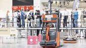 A robotized forklift truck on a demonstration area in an exhibition hall, in front of spectators on a platform.