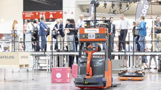 A robotized forklift truck on a demonstration area in an exhibition hall, in front of spectators on a platform.