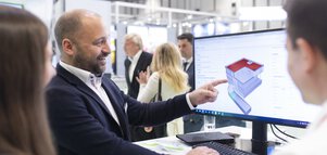 An exhibitor shows two visitors a model of a house on a computer screen and points to a detail.