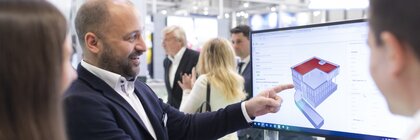 Ein Aussteller zeigt zwei Besuchern auf einen Computerbildschirm ein Modell eines Hauses und deutet dabei auf ein Detail.