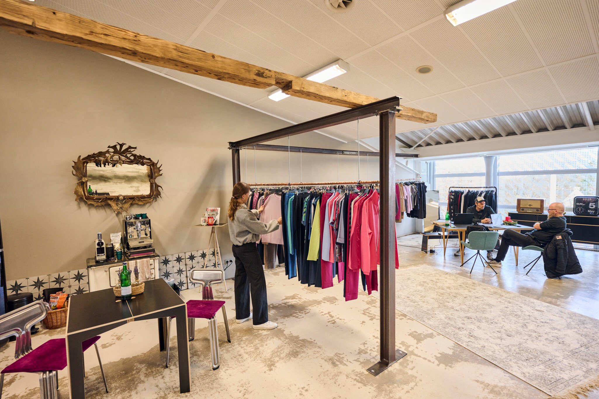 Ein modern eingerichteter Ausstellungsbereich für bunte Oberteile, die an Stahlträgern hängen. Im Hintergrund unterhalten sich zwei Männer an einem Tisch.