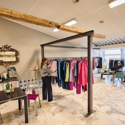 Ein modern eingerichteter Ausstellungsbereich für bunte Oberteile, die an Stahlträgern hängen. Im Hintergrund unterhalten sich zwei Männer an einem Tisch.