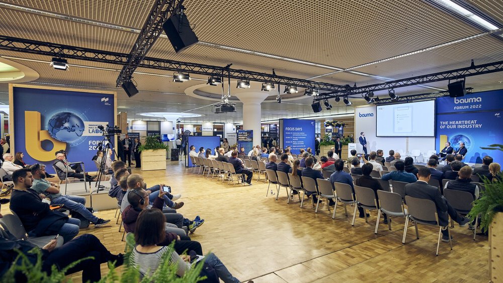 Audience sitting in a conference room listening to a speaker at the Bauma Forum 2022, with banners and screens displayed.