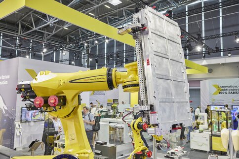 A yellow industrial robot arm lifts a large metal plate at a technology exhibition, with people visible in the background.