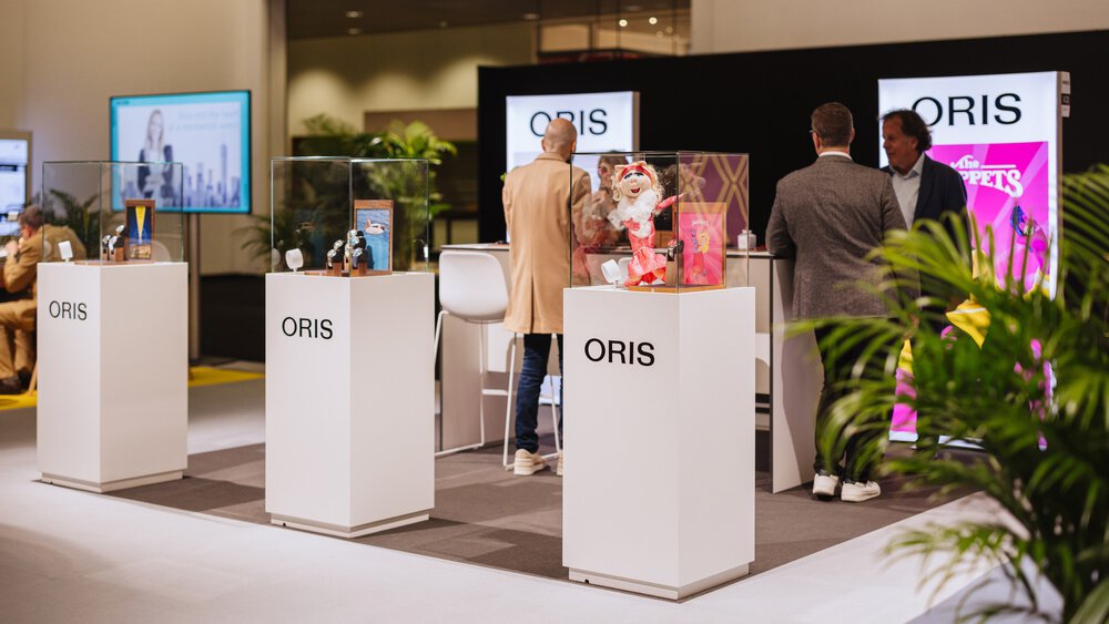 Three people stand at an ORIS stand with various articles and colourful posters behind glass display cases.