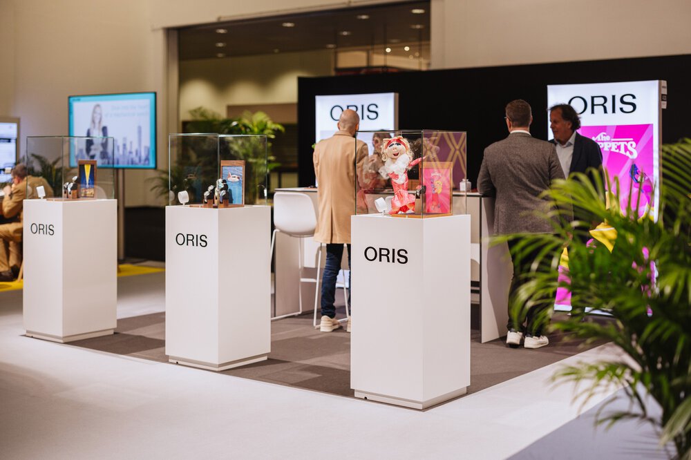 Drei Personen stehen an einem ORIS-Stand mit verschiedenen Artikeln und bunten Postern hinter Glasvitrinen.