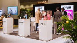 Drei Personen stehen an einem ORIS-Stand mit verschiedenen Artikeln und bunten Postern hinter Glasvitrinen.