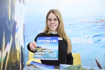 Lächelnde Frau hält an einem Messestand eine Broschüre zum Thema Seychellen in die Kamera. Im Hintergrund ist ein Werbeplakat.