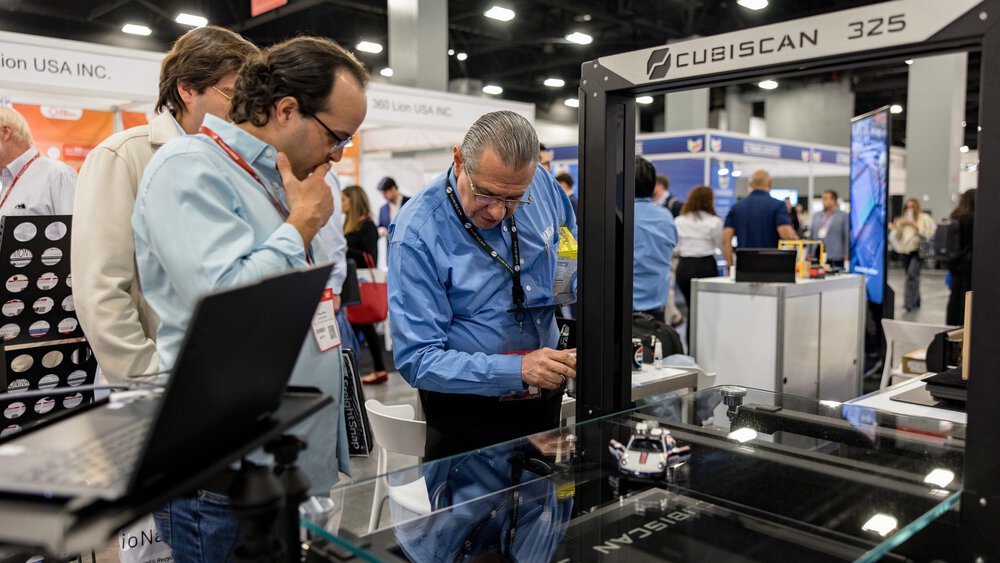Three men are standing in front of a large machine labelled “CUBISCAN 325” and looking at it with interest. Other trade fair visitors can be seen in the background.