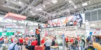 A well-attended exhibition stand displays various industrial equipment under bright lights and a large screen.