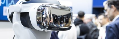 Eine weiße Schaufensterpuppe trägt ein futuristisches VR-Headset auf einer technischen Veranstaltung, während die Menschen im Hintergrund verschwimmen.