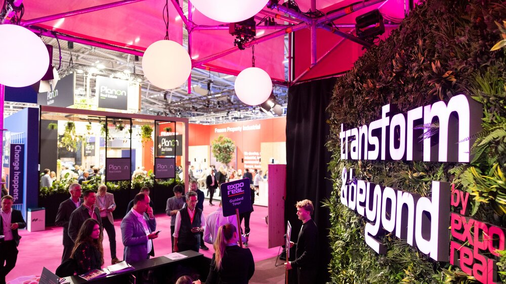 Messebereich mit Menschen an Ständen unter leuchtend pinker Beleuchtung und kugelförmigen Pendelleuchten, an einer mit Grünpflanzen geschmückten Wand prangt ein großes Transform & Beyond-Schild.