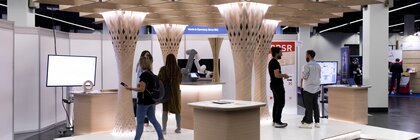 Futuristisches Standdesign mit Holzkonstruktionen und Roboterarm; Menschen stehen darin und unterhalten sich.