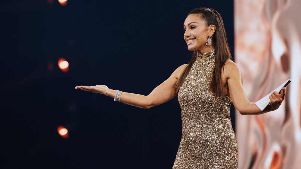 A woman with long brown hair in a glittering dress has her arms outstretched and is speaking to the audience.