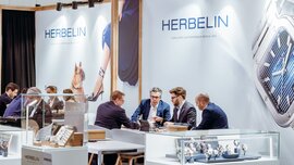 Ein Messestand mit Schmuckvitrinen und zahlreichen Personen in Anzügen, die sich, an Tischen sitzend, miteinander unterhalten.