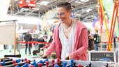 Eine lächelnde Person in einem rosafarbenen Kapuzenpulli spielt Tischfußball bei einer belebten Indoor-Veranstaltung oder Ausstellung.