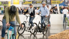Lächelnder Mann und lächelnde Frau probieren Fahrräder in einer Messehalle aus