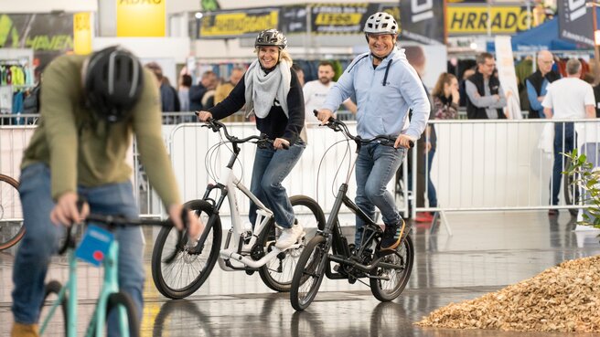 Lächelnder Mann und lächelnde Frau probieren Fahrräder in einer Messehalle aus