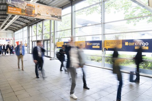Blurred silhouettes of trade fair visitors walk through a hall area with advertising signs to specific exhibition stands.