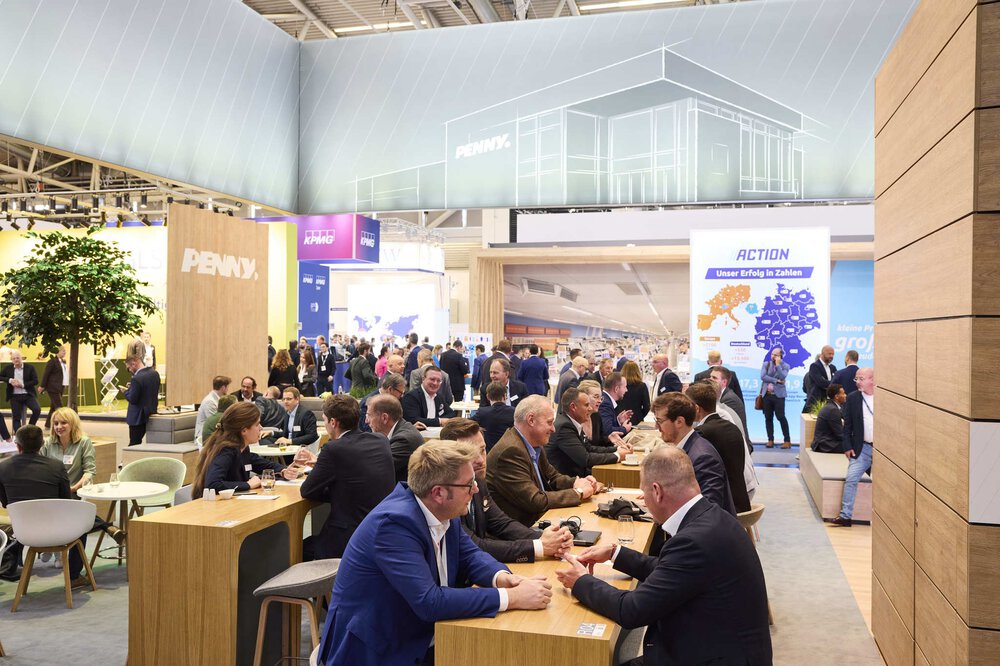 People sit and talk at tables at a large exhibition stand. Other stands can be seen in the background.