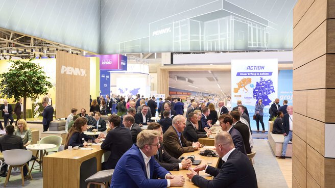 People sit and talk at tables at a large exhibition stand. Other stands can be seen in the background.