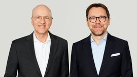 Zwei lächelnde Männer in Anzügen und mit Brillen stehen nebeneinander. Einer hat eine Glatze und der andere dunkle Haare und einen Bart.