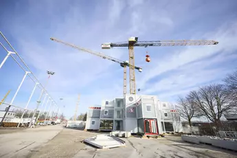 Zwei große Baukräne stehen vor einem Gebäude aus gestapelten Containern auf einer Baustelle. Einer der Kräne hebt eine orangefarbene Last. Der Himmel ist blau mit leichten Wolken, und links im Bild sind weitere Baugerüste und Beleuchtungsmasten zu sehen.