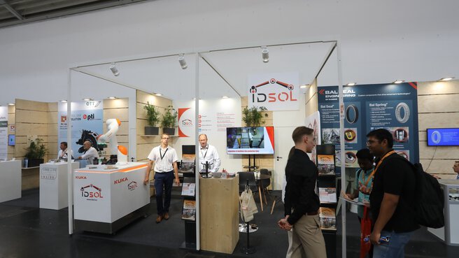 Trade fair booth featuring industrial robotics, product displays, and conversations between exhibitors and visitors in an exhibition hall.