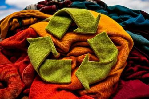 Ein grünes Recycling-Symbol aus Stoff liegt auf einem bunten Haufen verschiedener Textilien unter blauem Himmel. Das Bild symbolisiert Textilrecycling und Nachhaltigkeit.