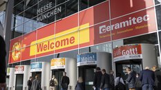 People walk past a trade fair entrance under a large sign with 