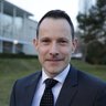 Ein Mann im Anzug mit gepunkteter Krawatte und kurzen dunklen Haaren lächelt in die Kamera.