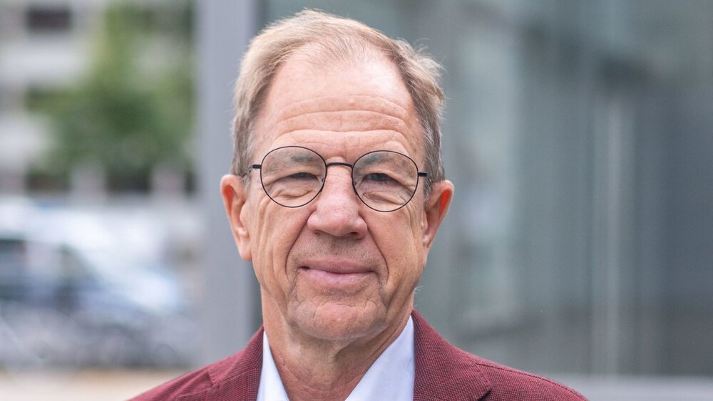 A smiling man with round glasses, a red jacket and a light-coloured shirt.