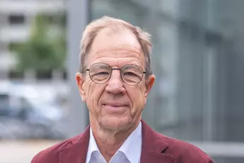 Ein lächelnder Mann mit runden Brillengläsern, rotem Jackett und hellem Hemd.