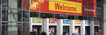 People enter the trade fair entrance with multilingual welcome signs on a glass façade.