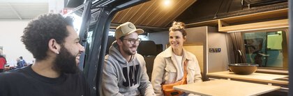 Zwei Personen sitzen lachend in einem Camper Van. Draußen steht eine dritte Person und schaut in den Van zu den Personen.