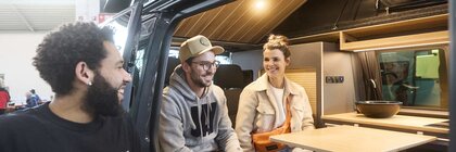 Zwei Personen sitzen lachend in einem Camper Van. Draußen steht eine dritte Person und schaut in den Van zu den Personen.