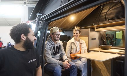 Zwei Personen sitzen lachend in einem Camper Van. Draußen steht eine dritte Person und schaut in den Van zu den Personen.
